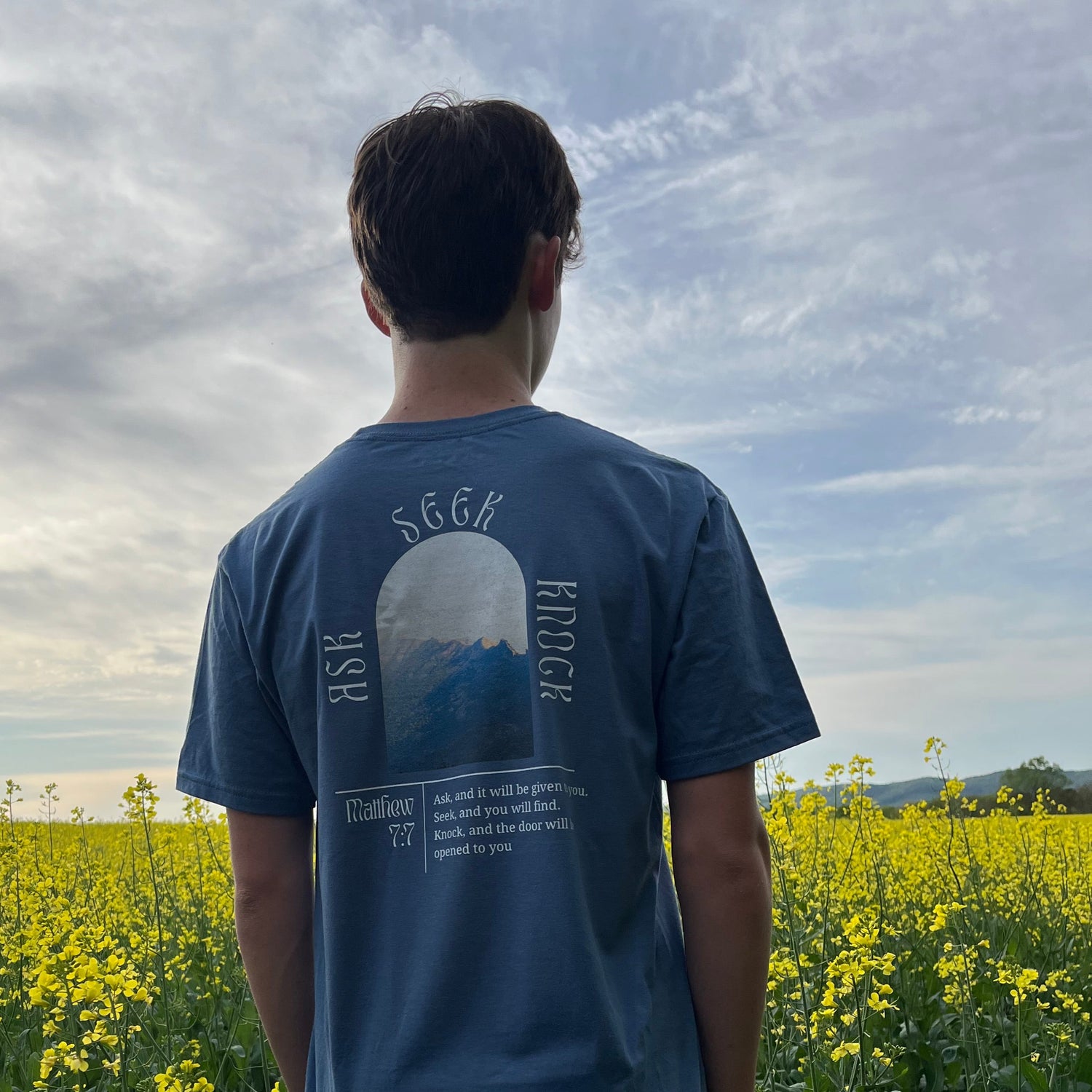 Person wearing a blue t-shirt with text standing in a field of yellow flowers.