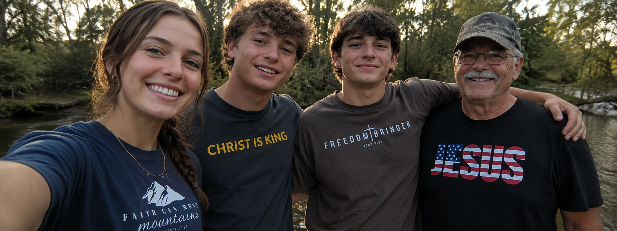 Four people posing together outdoors near water at sunset, wearing graphic t-shirts. The shirts feature the words 'CHRIST IS KING', 'FREEDOM BRIDGED', and 'JESUS' with an American flag pattern, alongside a white mountain logo on another shirt.