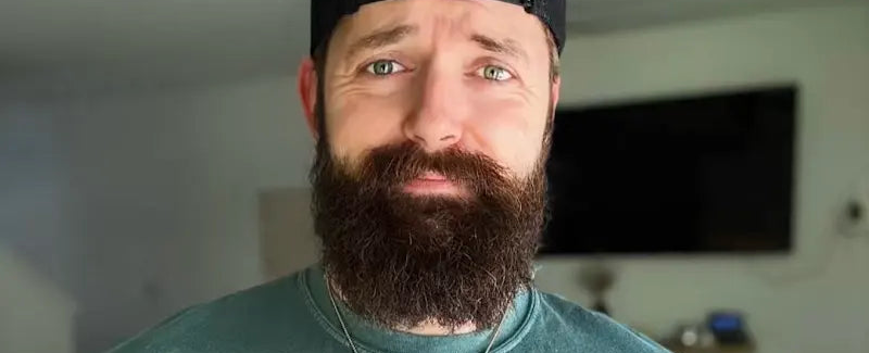 Man with a beard wearing a green t-shirt with a logo and a black cap indoors.